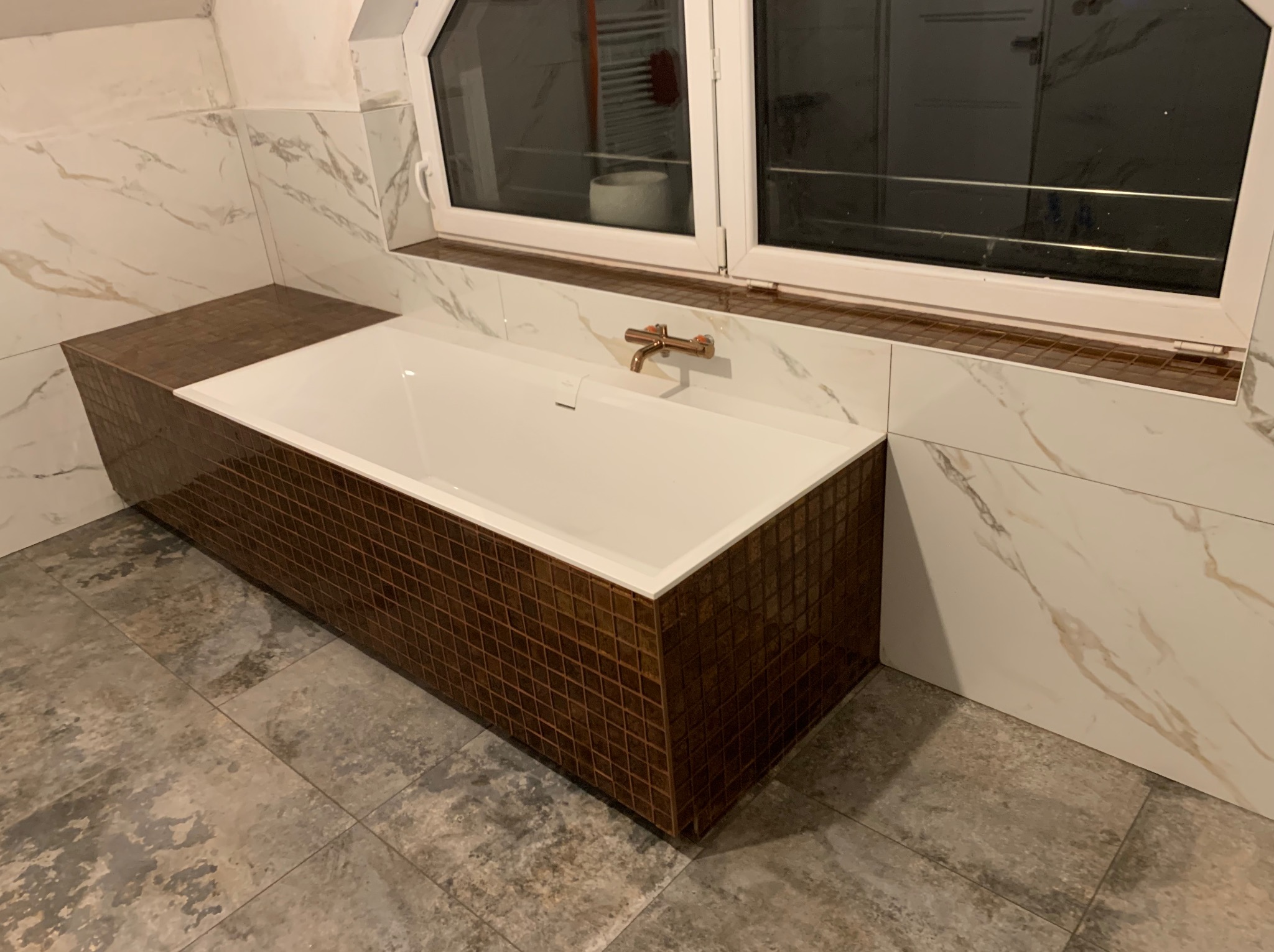 Biała wanna obudowana brązową mozaiką w łazience z marmurowymi płytkami i szarą podłogą. Widoczna bateria w kolorze miedzi i okno.