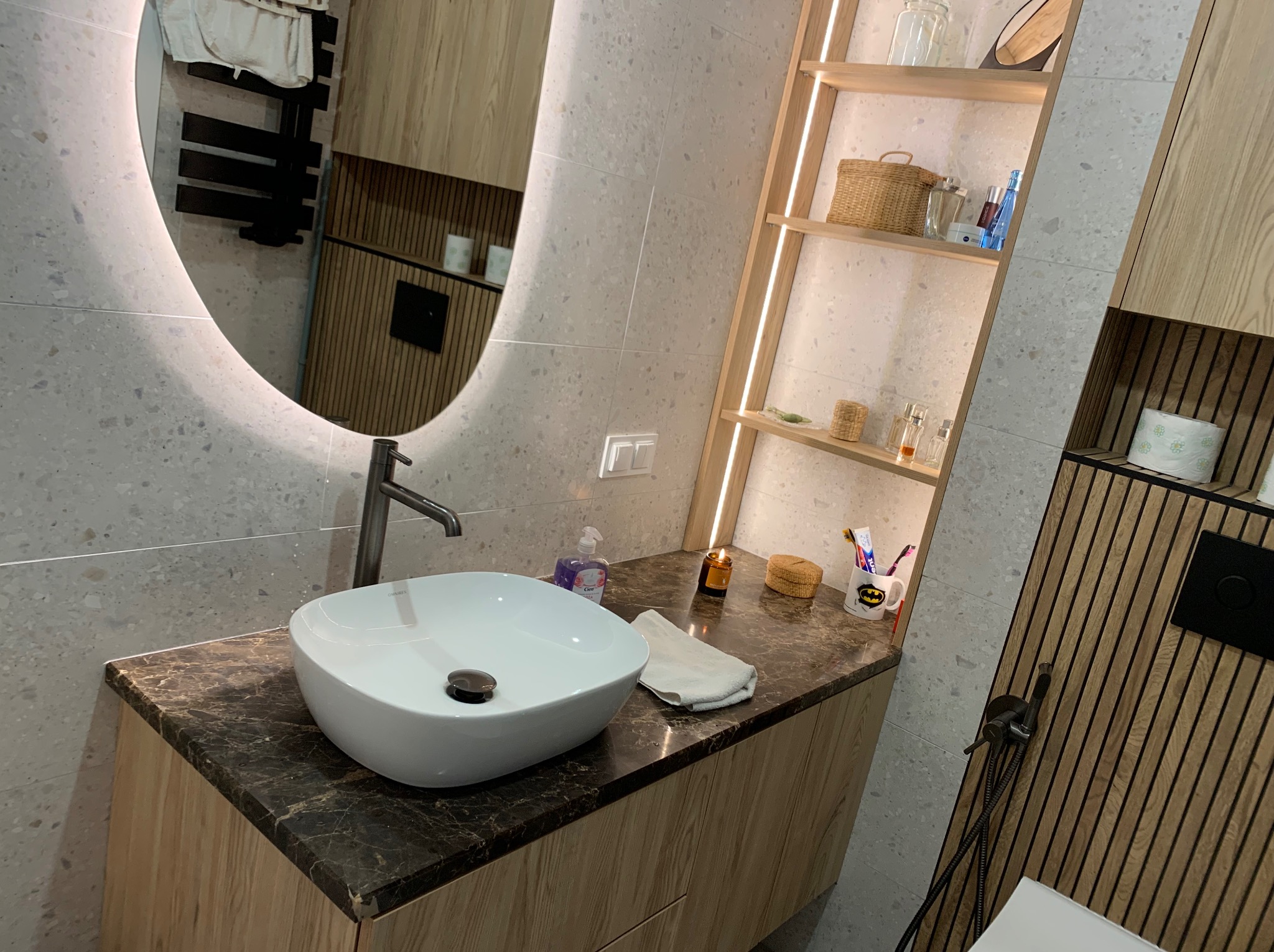 Nowoczesna łazienka z okrągłym lustrem, umywalką nablatową na blacie z ciemnego marmuru i drewnianymi półkami z oświetleniem LED.