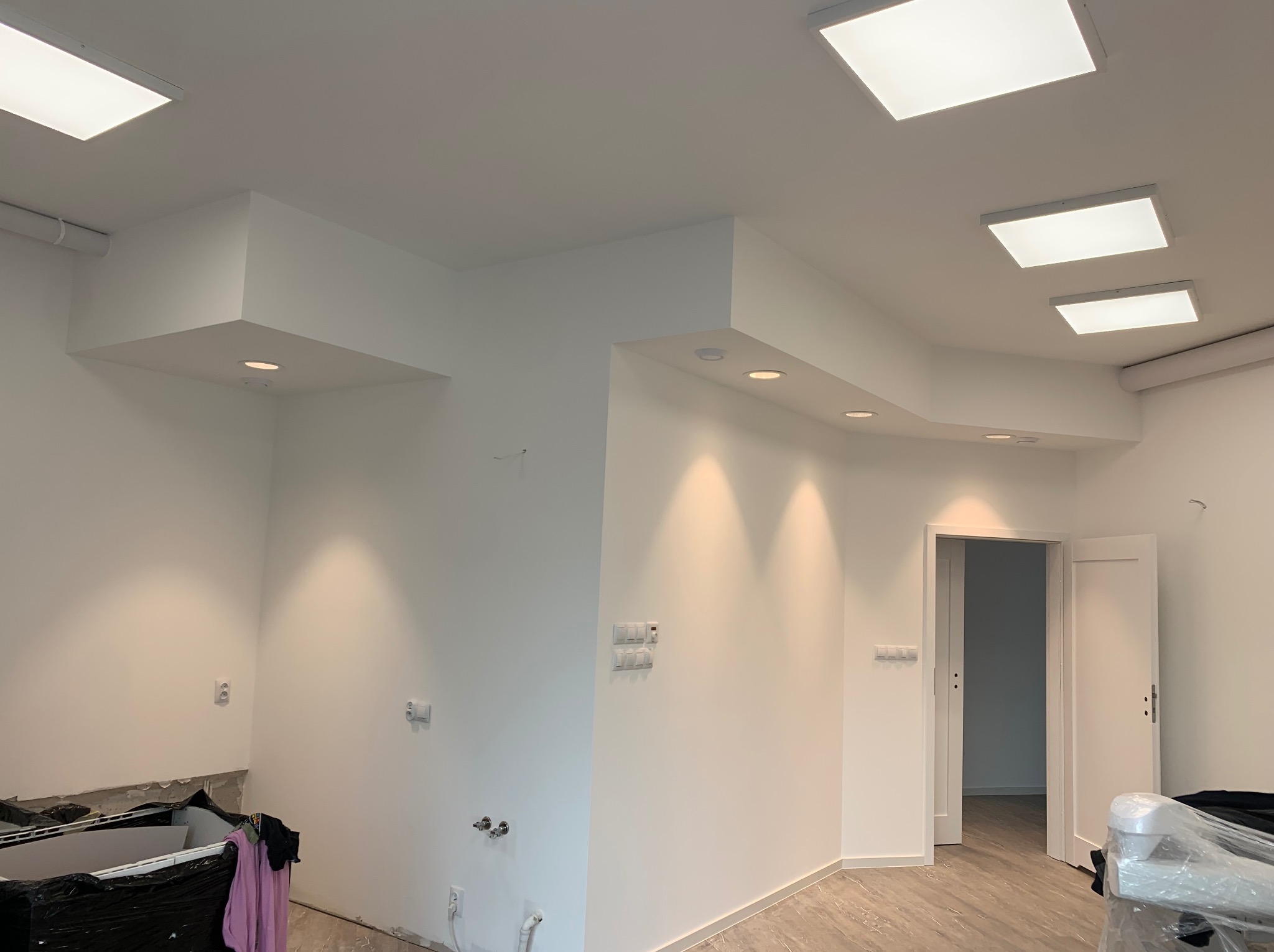 Białe wnętrze po malowaniu ścian, z widocznymi punktami świetlnymi w suficie, częściowo zabezpieczone podłoże, otwarte białe drzwi w tle i marmurowe płytki podłogowe.