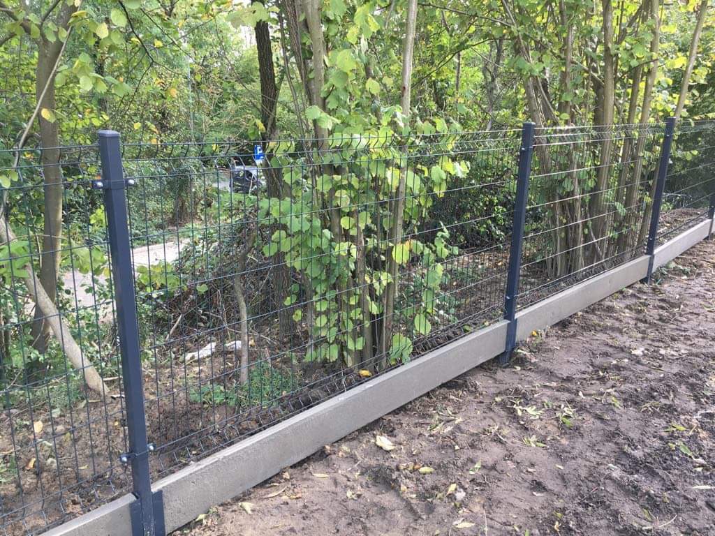 Nowoczesne ogrodzenie panelowe z siatki z podmurówką betonową, widoczne na tle zieleni drzew i krzewów.