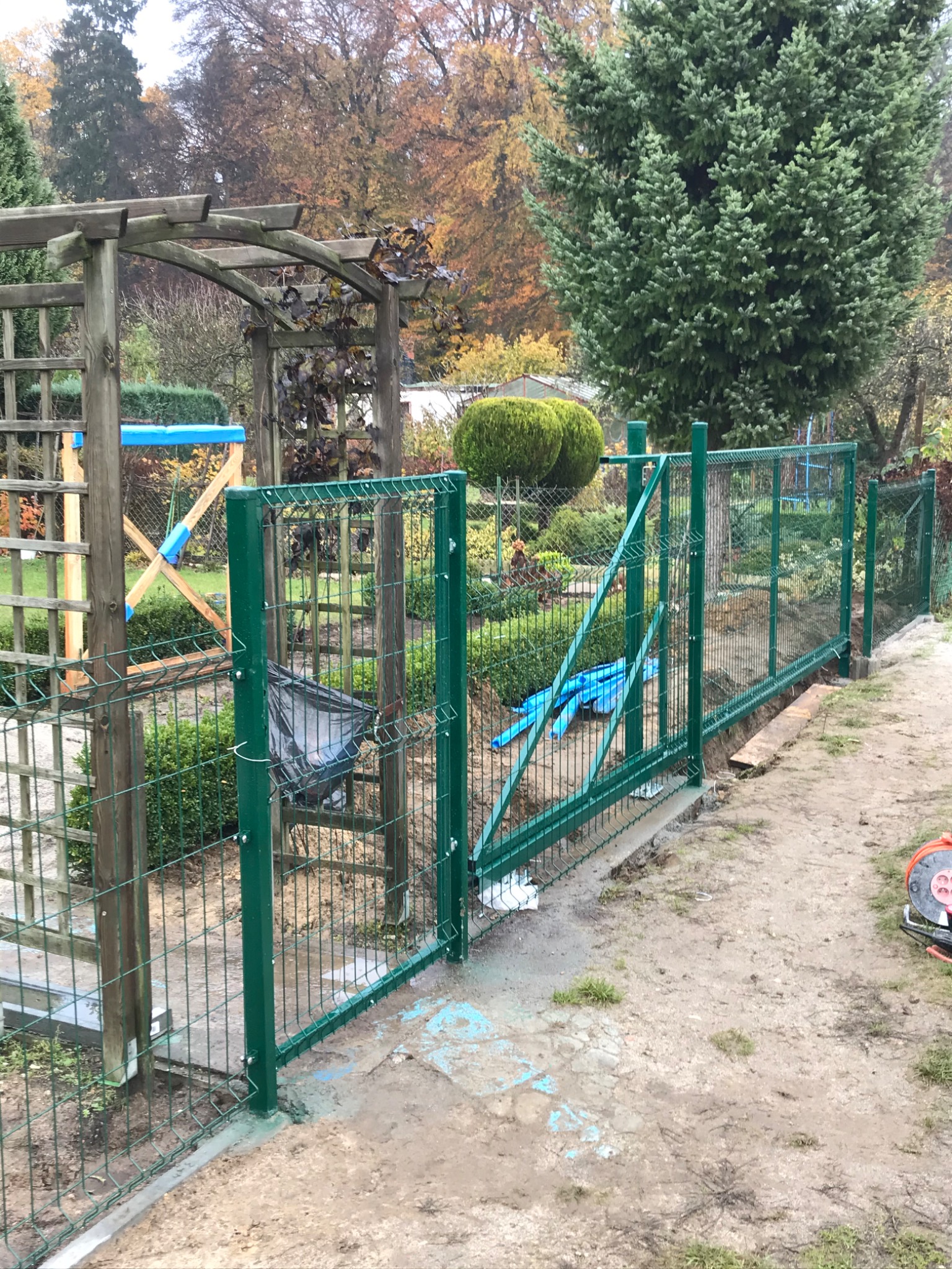 Zielone ogrodzenie panelowe z furtką w trakcie montażu na działce, widoczne elementy betonowe fundamentu i częściowo ułożona siatka, w tle pergola i drzewa.