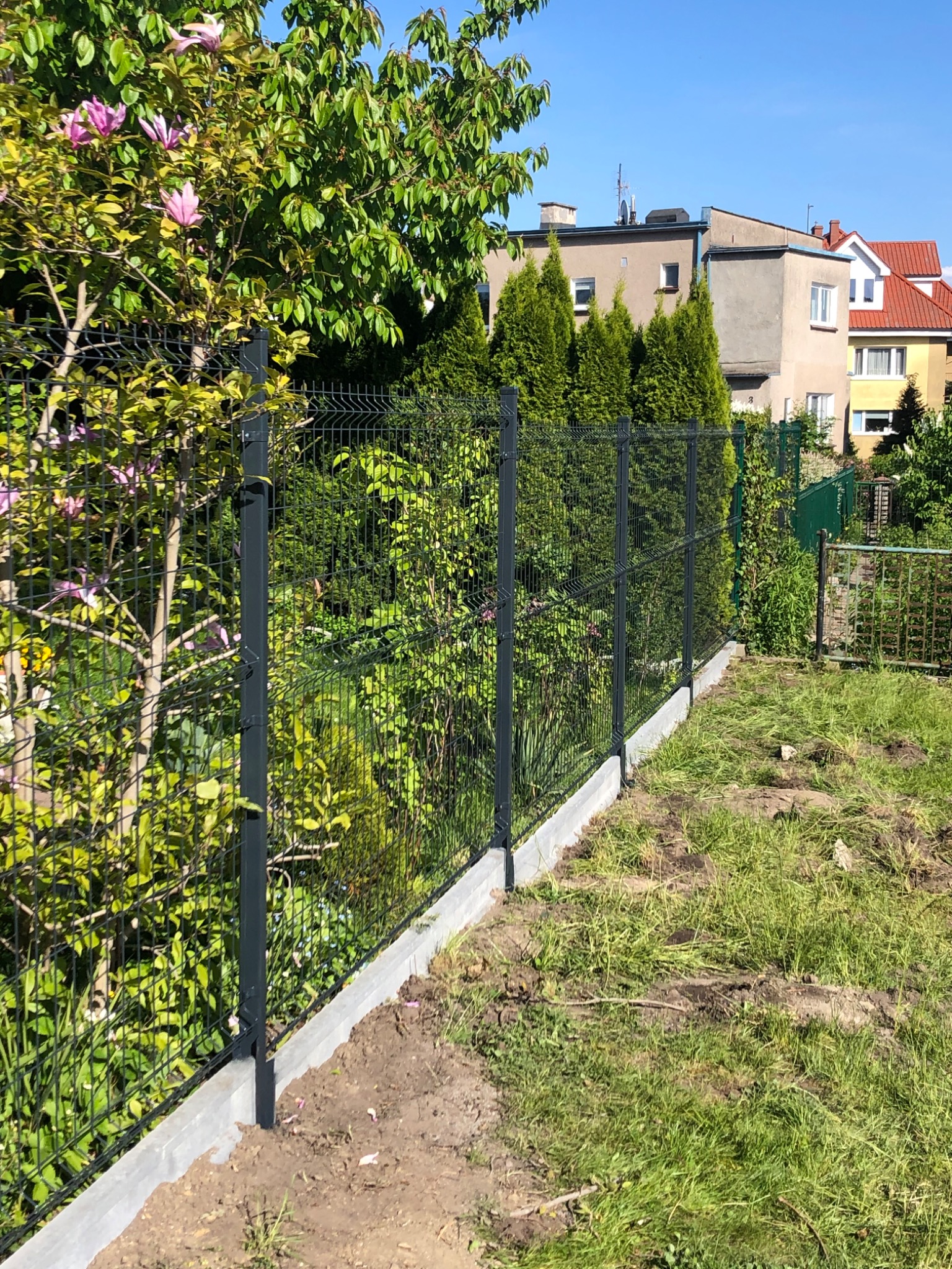 Nowoczesne, antracytowe ogrodzenie panelowe z podmurówką betonową, widok z perspektywy trawnika z młodą trawą, w tle zieleń drzew i zabudowa jednorodzinna.