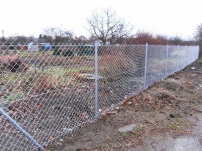 Długie, metalowe ogrodzenie z siatki, z widocznymi słupkami, otaczające teren z niską roślinnością, na tle drzew i zabudowań w pochmurny dzień.