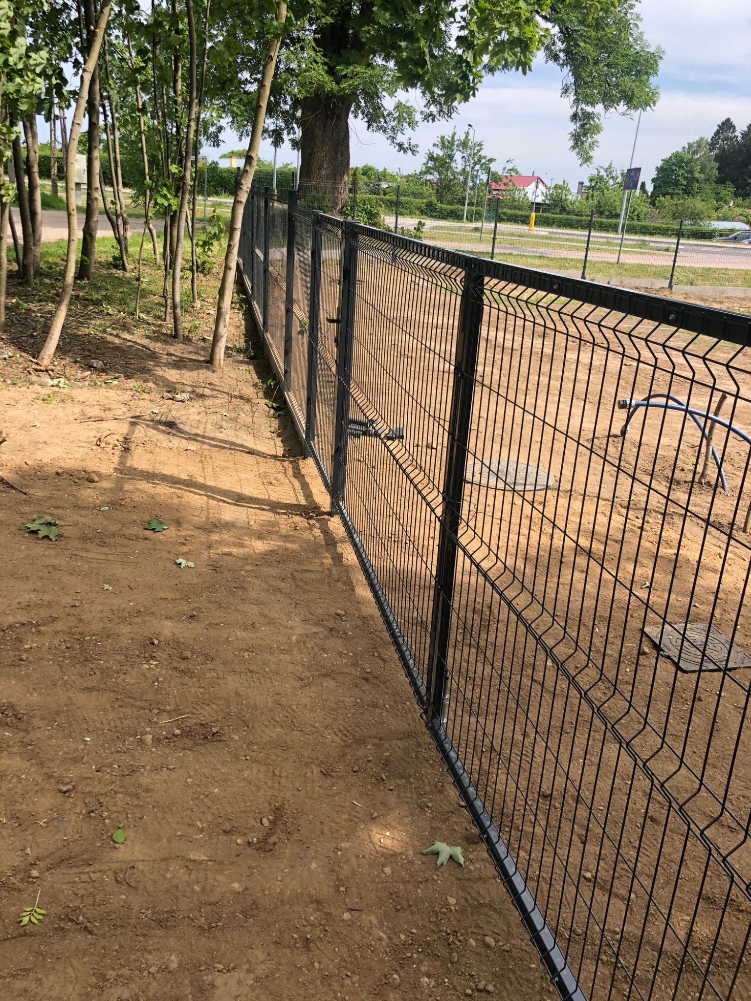 Czarny panelowy płot z furtką, widoczny z perspektywy gruntu, z drzewami w tle i śladami opon na ziemi.