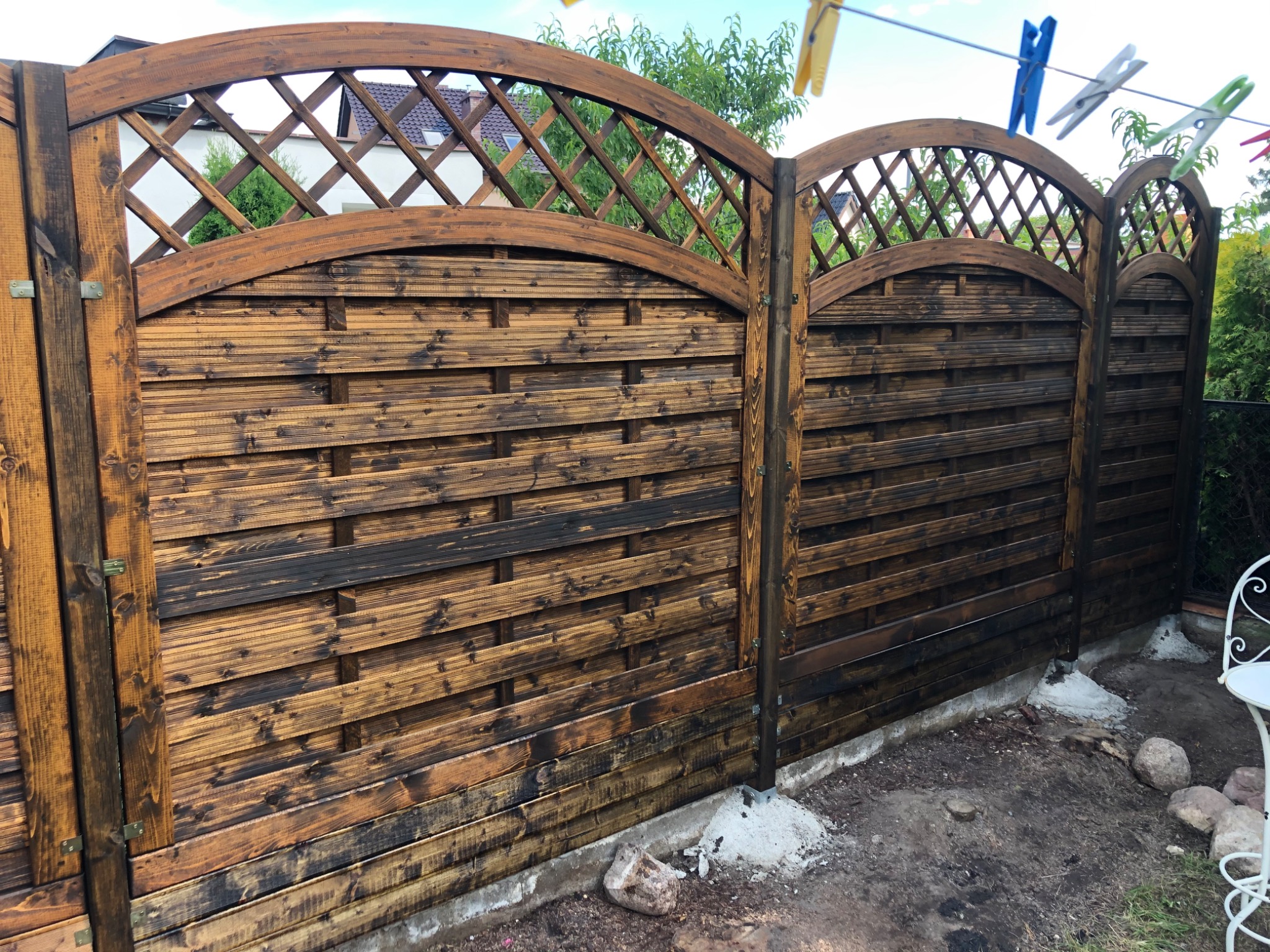 Drewniane ogrodzenie z łukowym wykończeniem i kratką, świeżo zaimpregnowane, z widocznymi śladami impregnatu, stojące na betonowej podmurówce, z ubraniami suszącymi się na sznurku w tle.