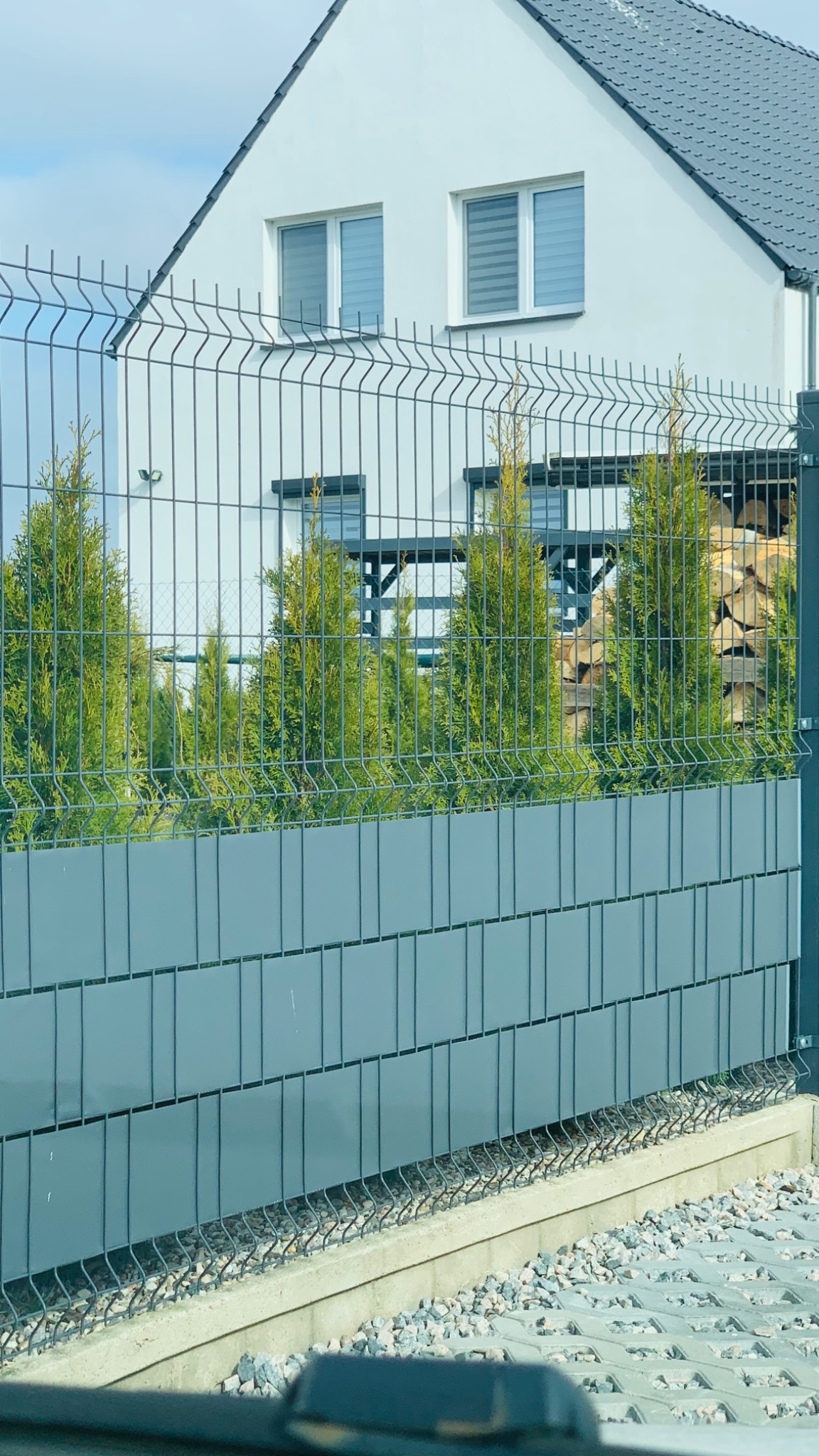 Panelowa, metalowa siatka ogrodzeniowa z podmurówką i przesłoną z szarych paneli, za nią rząd iglaków i stos drewna na tle białego domu z szarym dachem.