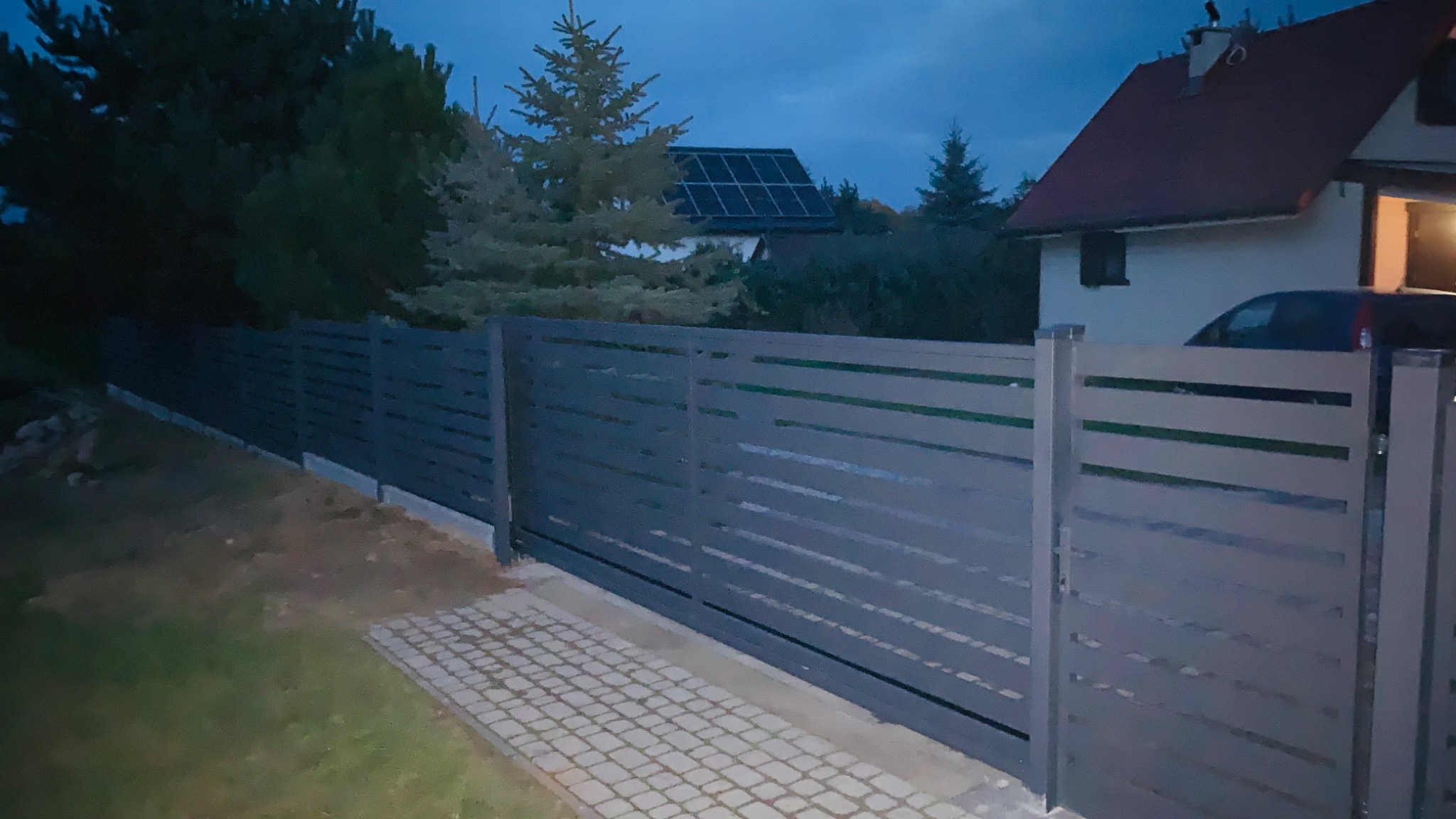 Nowoczesne, poziome ogrodzenie panelowe w odcieniach szarości, z przesuwną bramą wjazdową, na tle domu z panelami słonecznymi na dachu, sfotografowane o zmierzchu.