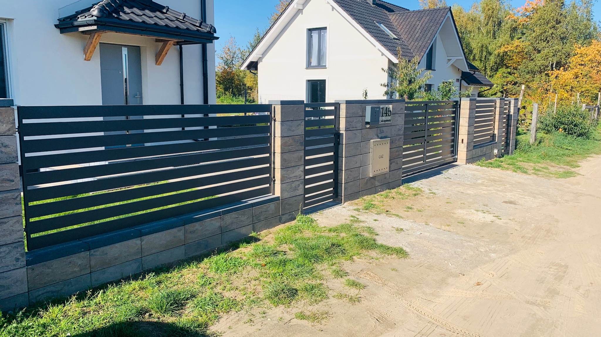 Nowoczesne ogrodzenie panelowe z poziomymi przęsłami w kolorze grafitowym, osadzone na słupkach z szarego kamienia łupanego, z furtką i skrzynką na listy przy numerze posesji 14b, na tle dwóch...