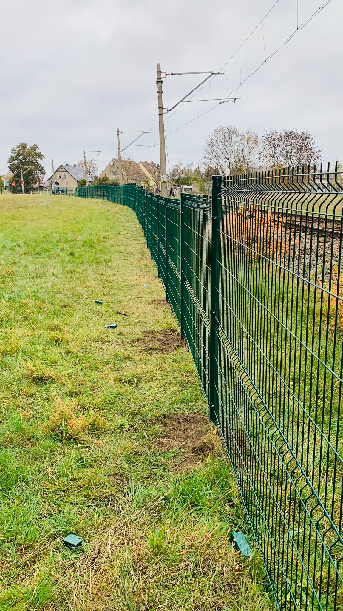 Długie, zielone ogrodzenie z siatki panelowej, biegnące wzdłuż torów kolejowych na tle pochmurnego nieba i zabudowań mieszkalnych. Widoczne plastikowe zaślepki na słupkach ogrodzeniowych.