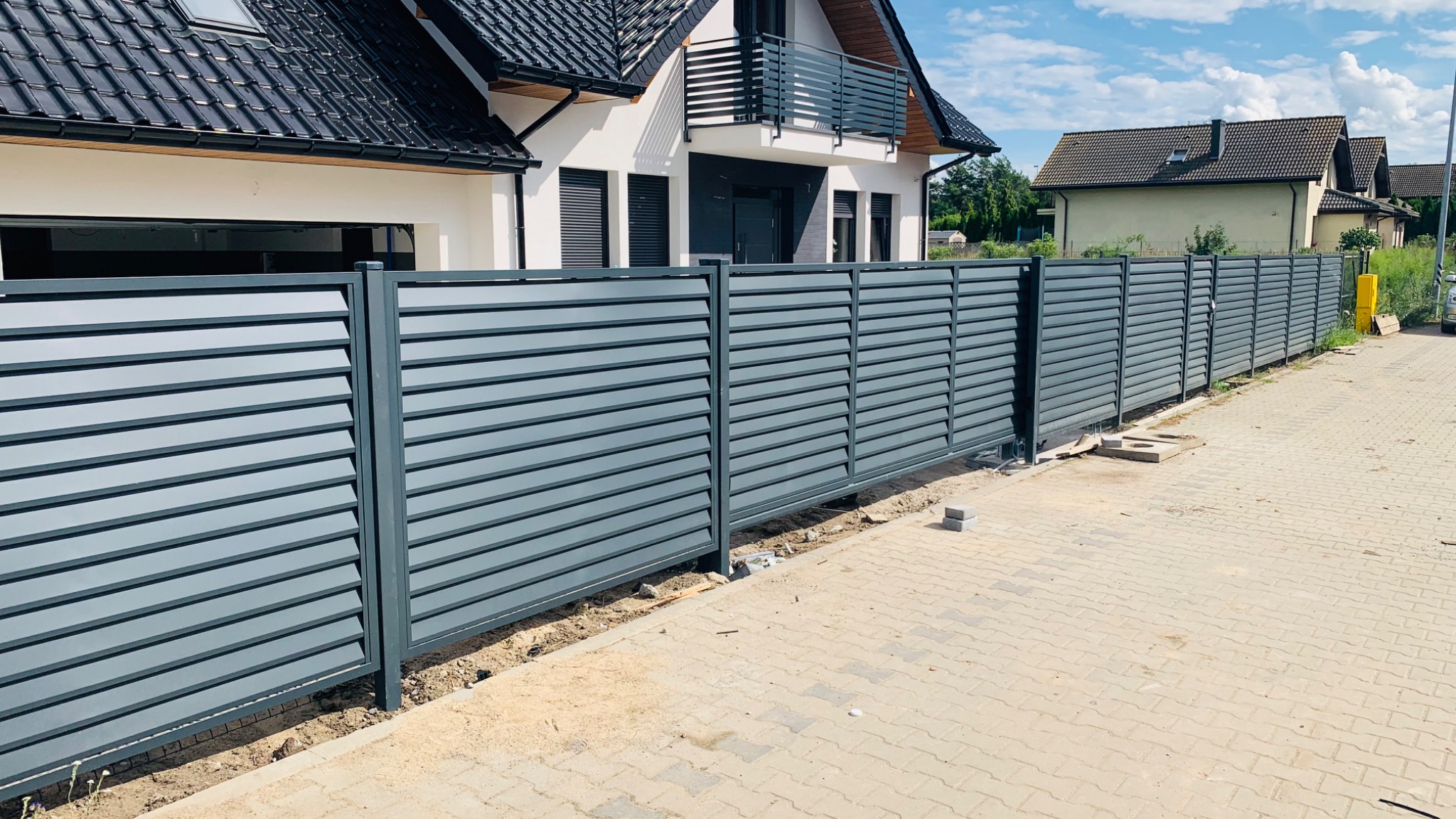 Nowoczesne, poziome, szare ogrodzenie panelowe z metalu, widok z zewnątrz posesji, na pierwszym planie kostka brukowa, w tle dom jednorodzinny z balkonem.