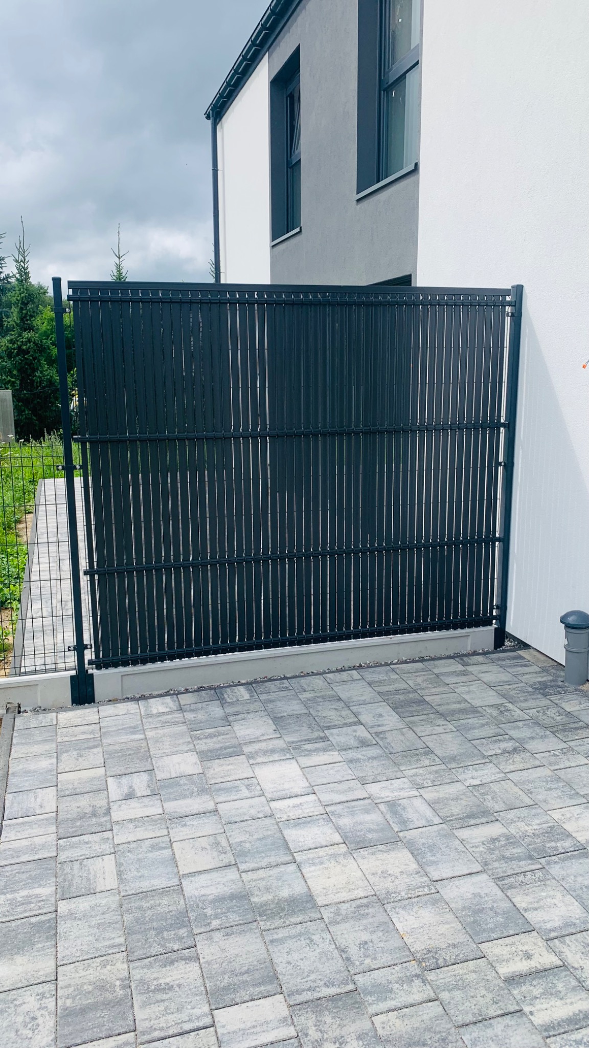 Nowoczesne, antracytowe ogrodzenie panelowe z pionowych listew, oddzielające posesję od siatki ogrodzeniowej, na tle dwukondygnacyjnego domu z szarą elewacją i kostki brukowej.