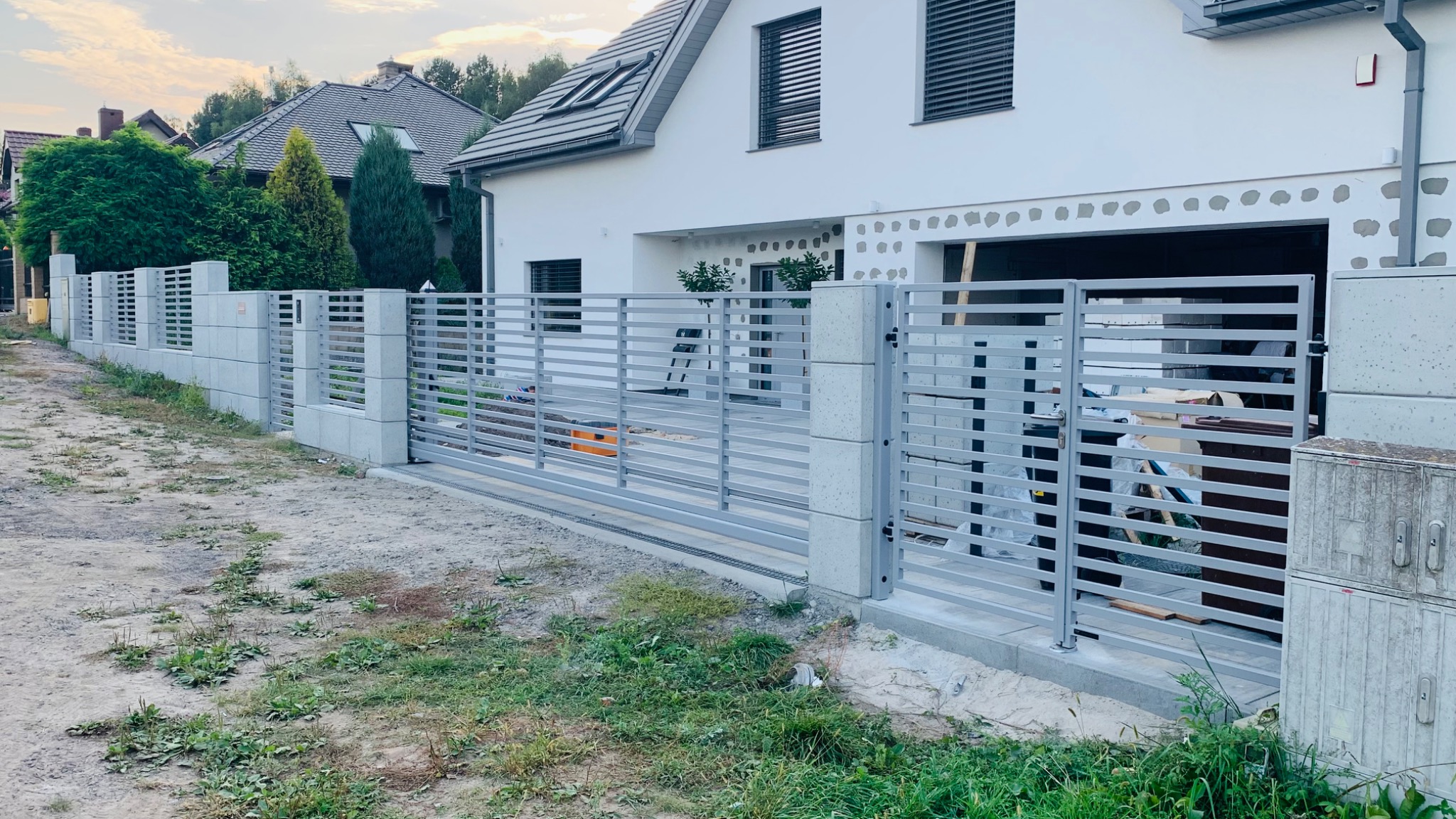 Nowoczesne ogrodzenie z szarej stali i betonowych słupków przed białym domem z szarą dachówką, widoczne elementy montażowe i teren po budowie.