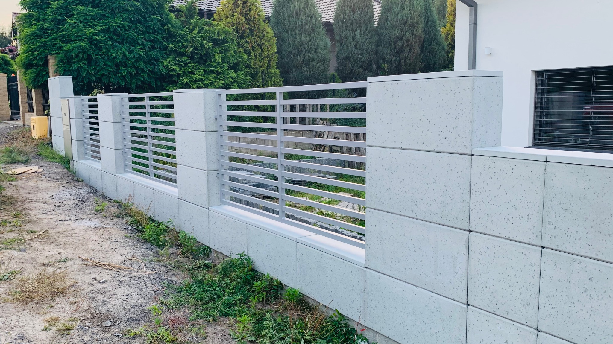Nowoczesne ogrodzenie z betonowych bloczków i poziomych, metalowych przęseł, przylegające do białego budynku z oknem z żaluzjami.