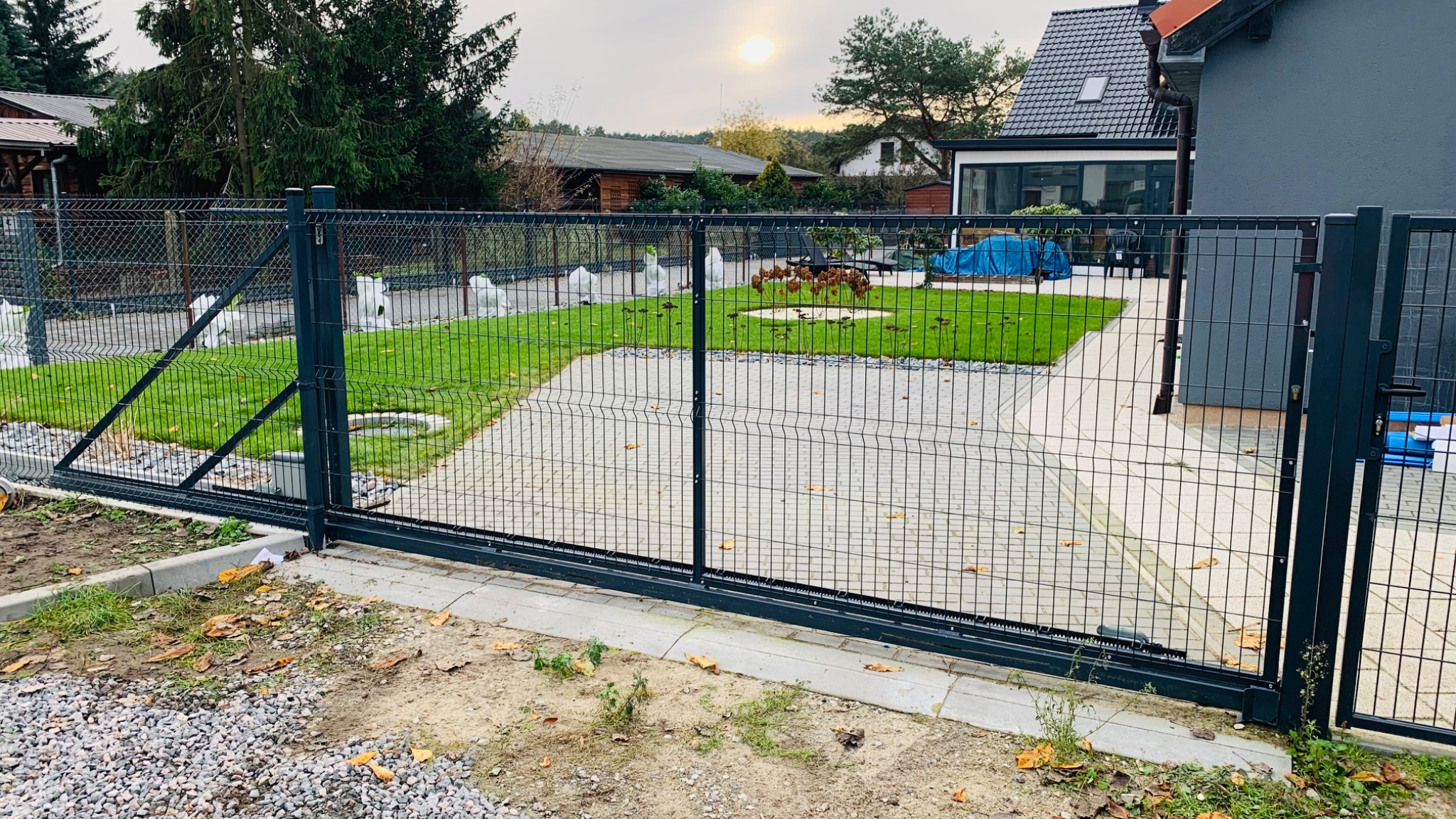 Przesuwna brama z siatki panelowej w kolorze antracytowym, widok od strony podjazdu wyłożonego kostką, trawnik z elementami dekoracyjnymi w tle, pochmurne niebo.