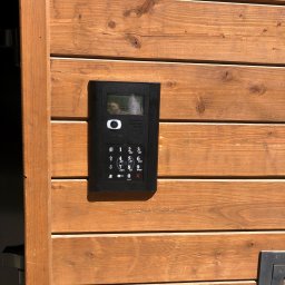 NapadSystem Mirosław Skałka - Czarny panel wideodomofonu z klawiaturą i wyświetlaczem zamontowany na drewnianej bramie wjazdowej.