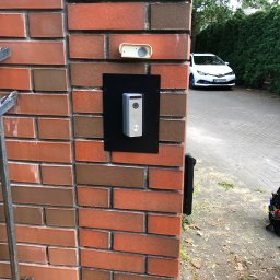 NapadSystem Mirosław Skałka - Zewnętrzny panel domofonowy z kamerą i przyciskiem dzwonka, zamontowany na czarnej płycie na ceglanym słupku ogrodzenia, widoczny biały samochód i torba z narzędziami.