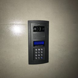 NapadSystem Mirosław Skałka - Zamontowany, czarny wideodomofon z kamerą i panelem numerycznym na jasnej, błyszczącej ścianie w Warszawie. Widoczny wyświetlacz z instrukcjami i przyciski numeryczne.