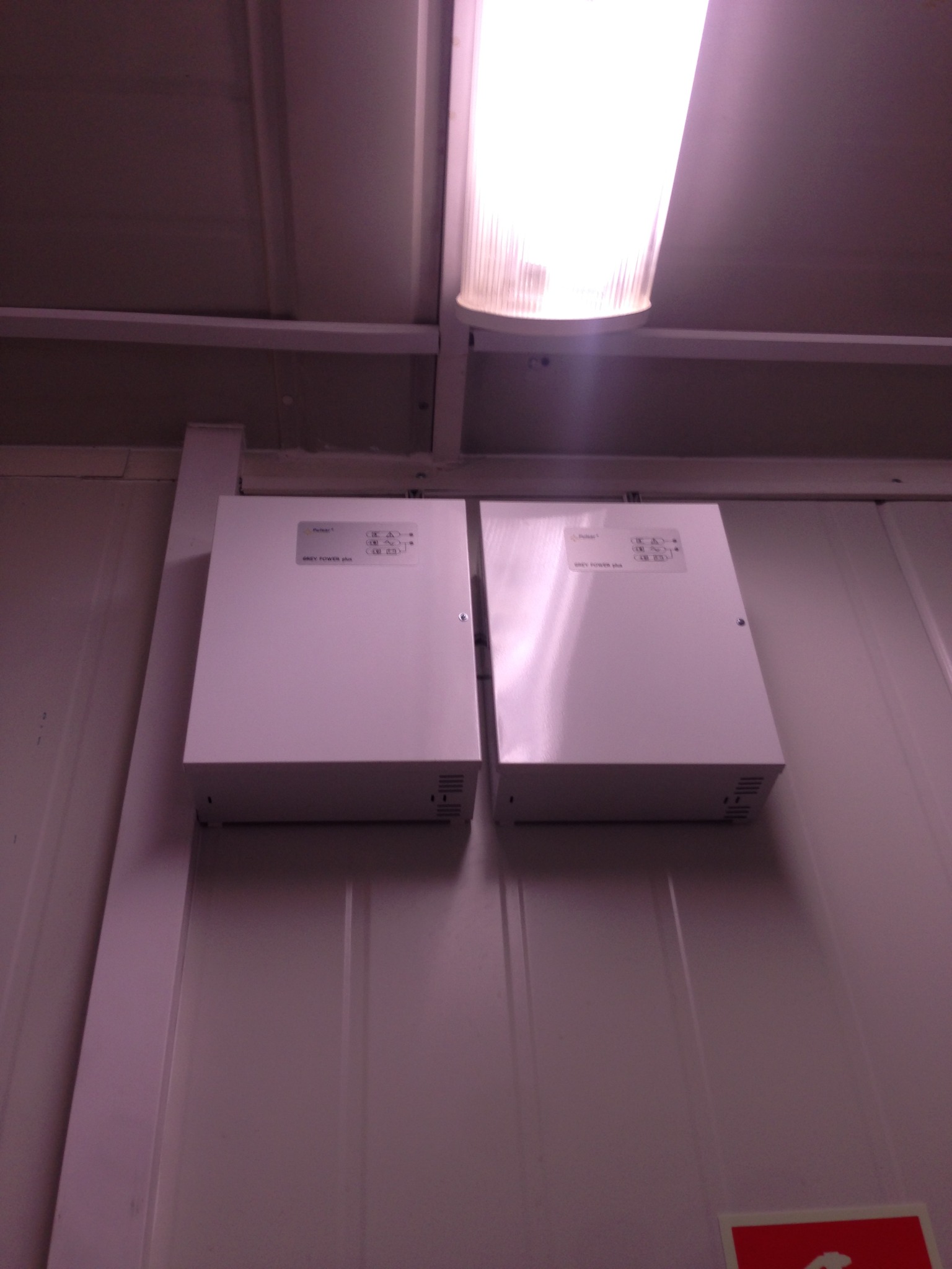 Dwa białe zasilacze impulsowe marki Pulsar Grey Power Plus zamontowane na białej ścianie, oświetlone jarzeniówką.