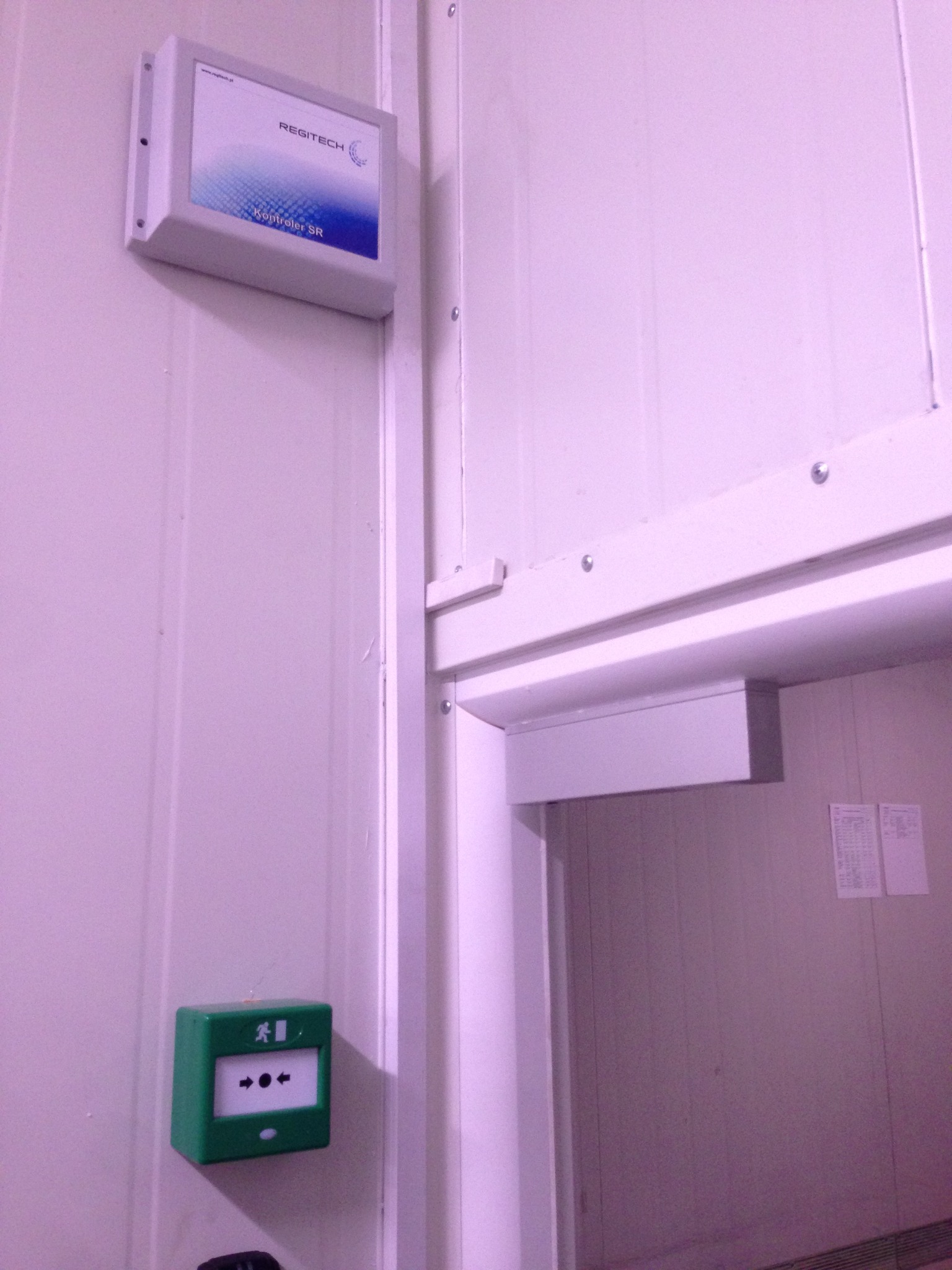 Zamontowany na ścianie kontroler Regitech Kontroler SR oraz zielony przycisk alarmowy kierujący do wyjścia ewakuacyjnego, widoczne przy wejściu do pomieszczenia.
