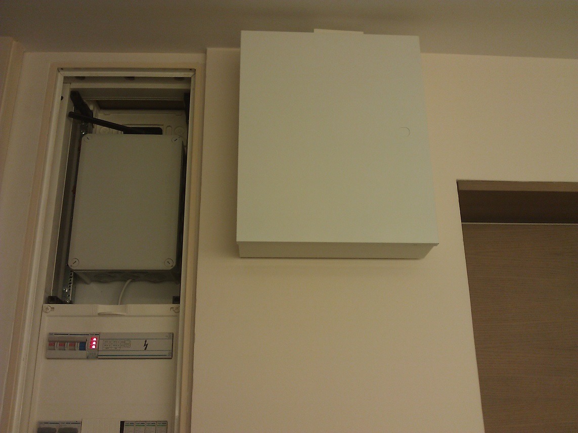 Zamknięta, biała skrzynka z bezpiecznikami i okablowaniem, wbudowana w ścianę obok drewnianych drzwi, widoczne elementy instalacji elektrycznej.