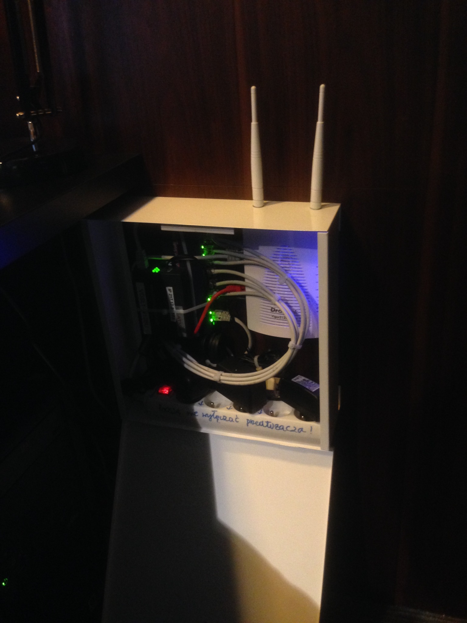 Otwarta biała szafka z routerem, przewodami i zasilaczami, widoczne diody LED oraz napis 'Proszę nie wyłączać przetwornika!'