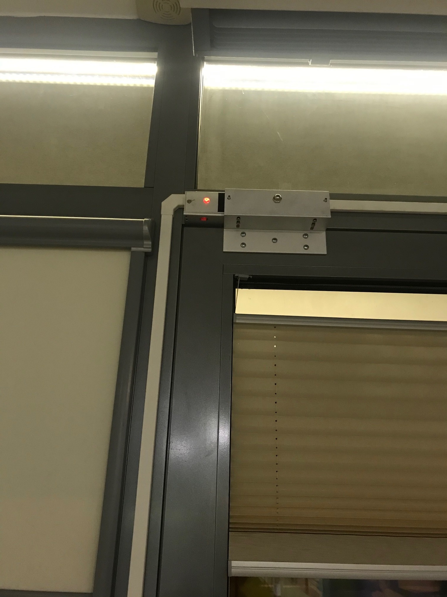 Elektromagnetyczny zamek drzwiowy z czerwoną diodą LED, zamontowany na szarej ramie drzwiowej w biurze, widoczne żaluzje w oknie.