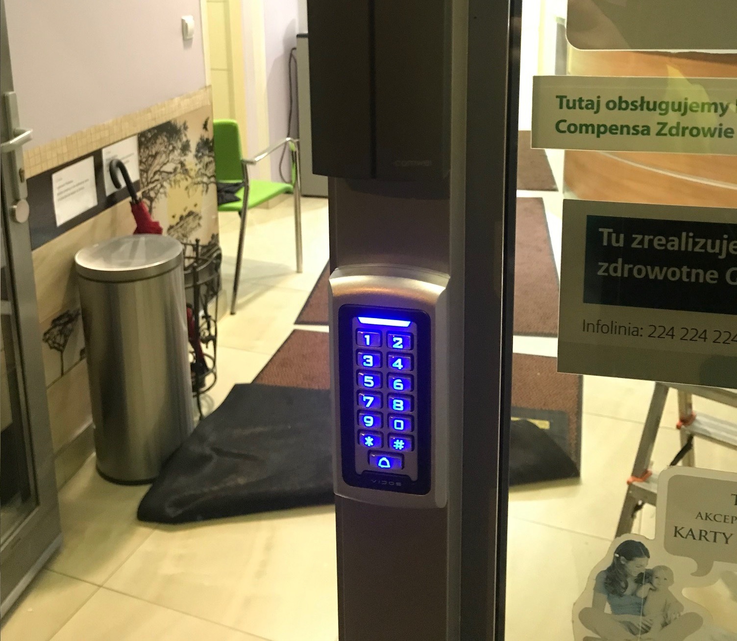 Klawiatura numeryczna domofonu Vidos z podświetlonymi na niebiesko przyciskami, zamontowana przy wejściu do placówki medycznej z widocznymi naklejkami informacyjnymi i metalowym koszem na śmieci...