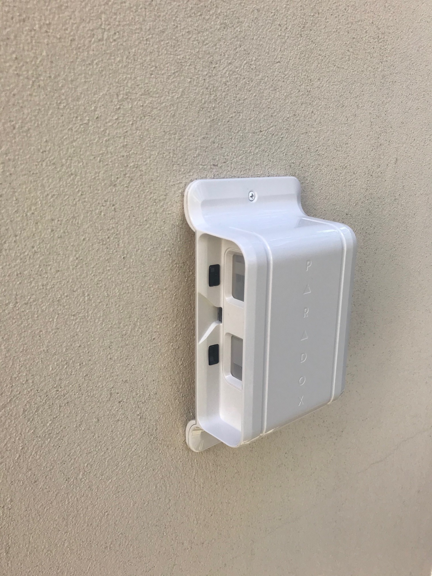 Biała obudowa czujnika alarmowego firmy Paradox zamontowana na beżowej, teksturowanej ścianie zewnętrznej budynku.