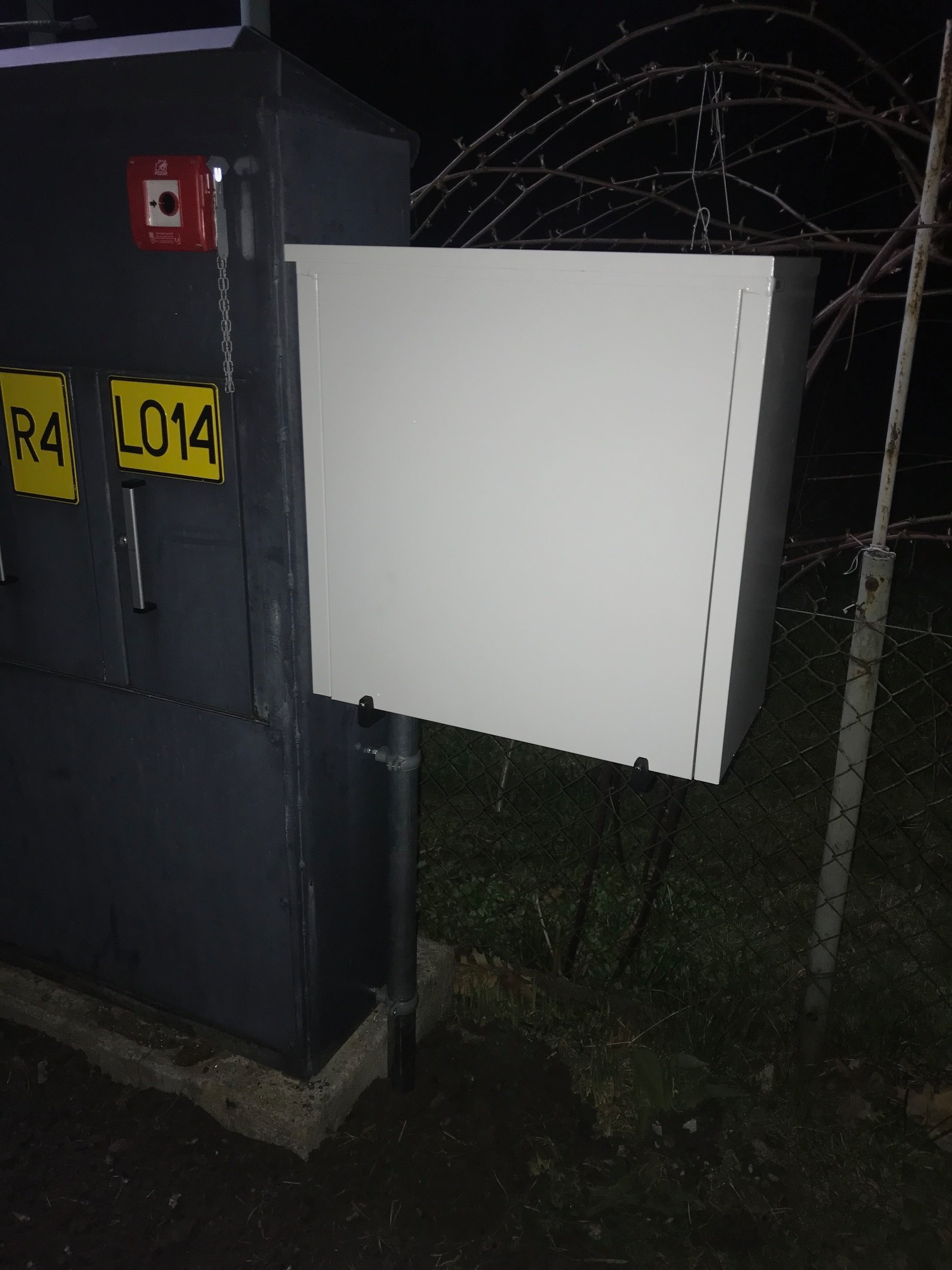 Zewnętrzna szafa sterownicza z zamontowanym panelem dostępowym, widoczny przycisk alarmowy na obudowie, oświetlenie nocne, metalowe ogrodzenie w tle.