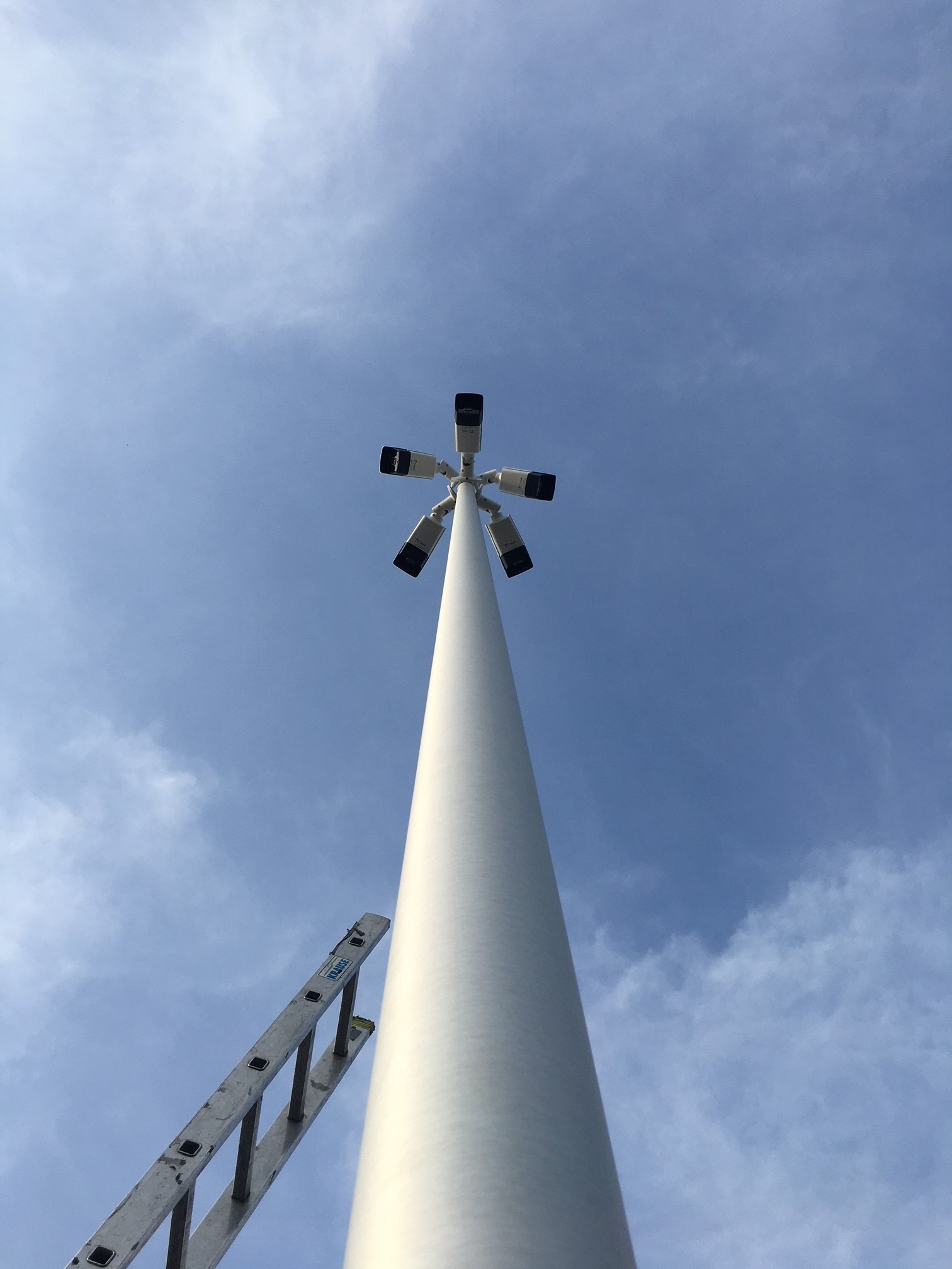 Wysoki maszt z pięcioma kamerami monitoringu, widziany z dołu, z częściowo widoczną drabiną opartą o maszt na tle błękitnego nieba z chmurami.