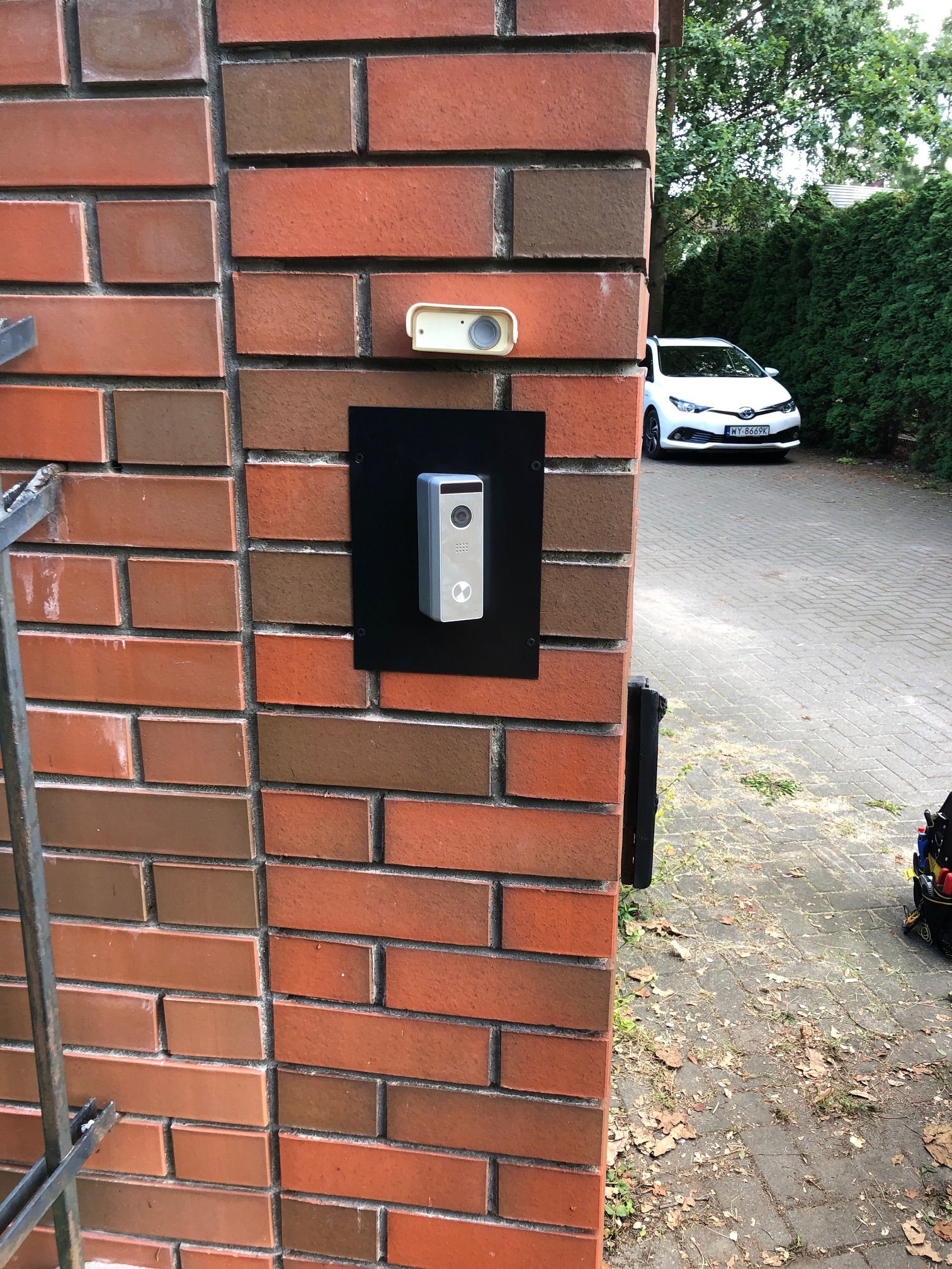 Zewnętrzny panel domofonowy z kamerą i przyciskiem dzwonka, zamontowany na czarnej płycie na ceglanym słupku ogrodzenia, widoczny biały samochód i torba z narzędziami.