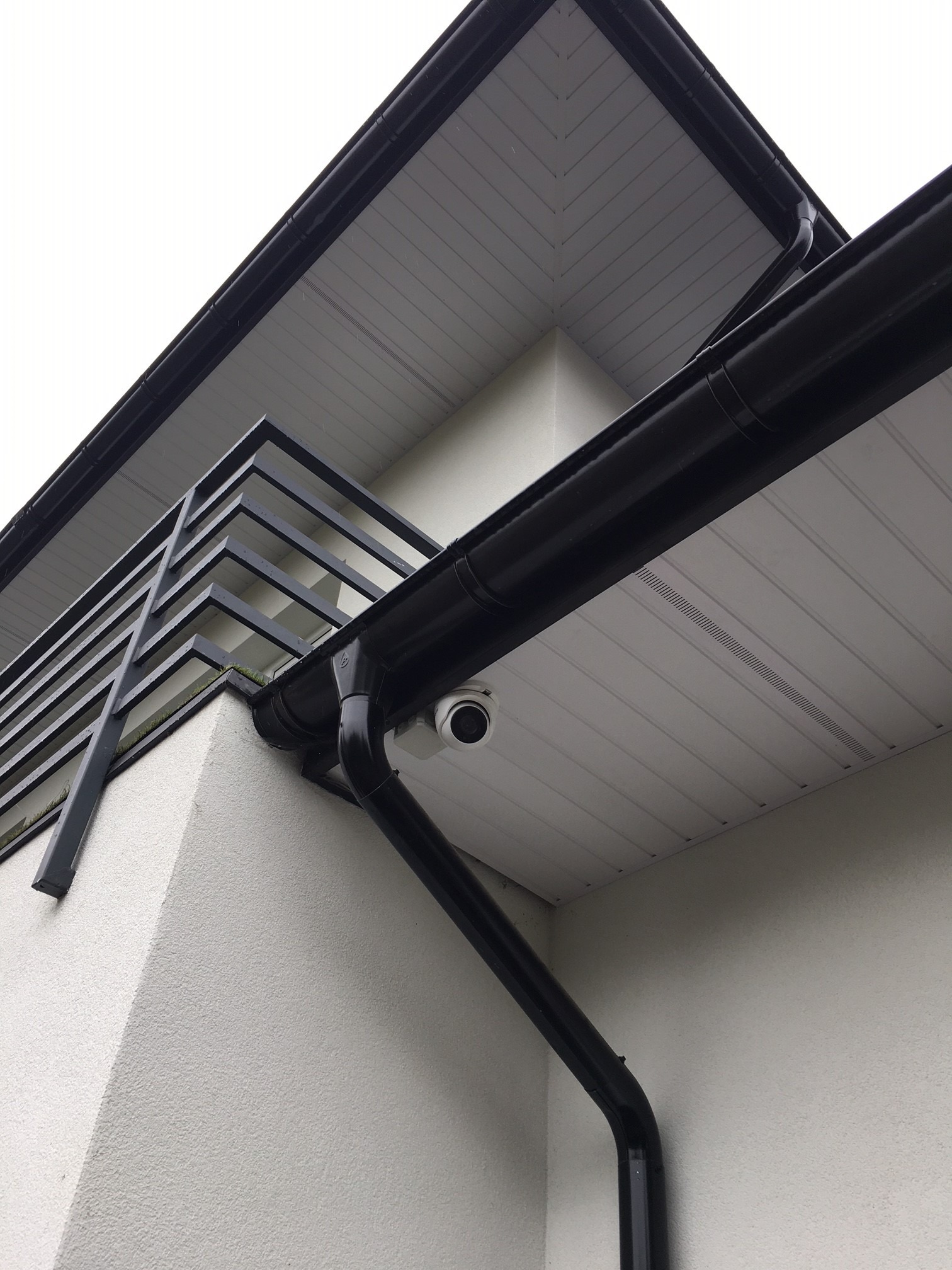 Kamera monitoringu pod okapem nowoczesnego domu z czarnymi rynnami, widok z dołu na elewację i balkon z metalową balustradą.