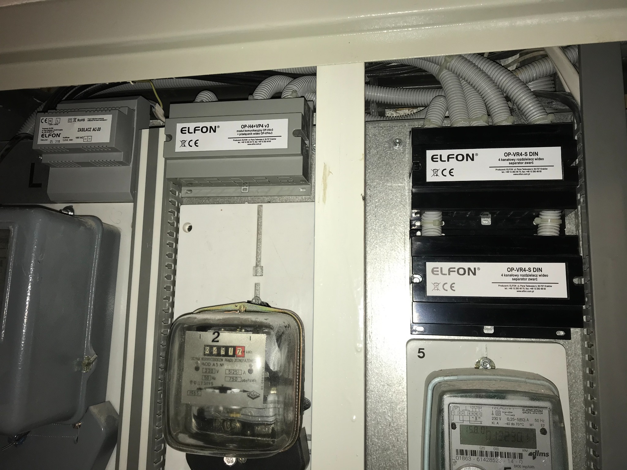 Szara szafa elektryczna z dwoma licznikami energii, zasilaczem AC-20, modułem komunikacyjnym OP-H4+VP4 v3 oraz dwoma rozdzielaczami wideo OP-VR4-S DIN firmy Elfon, widoczne przewody w peszlach.