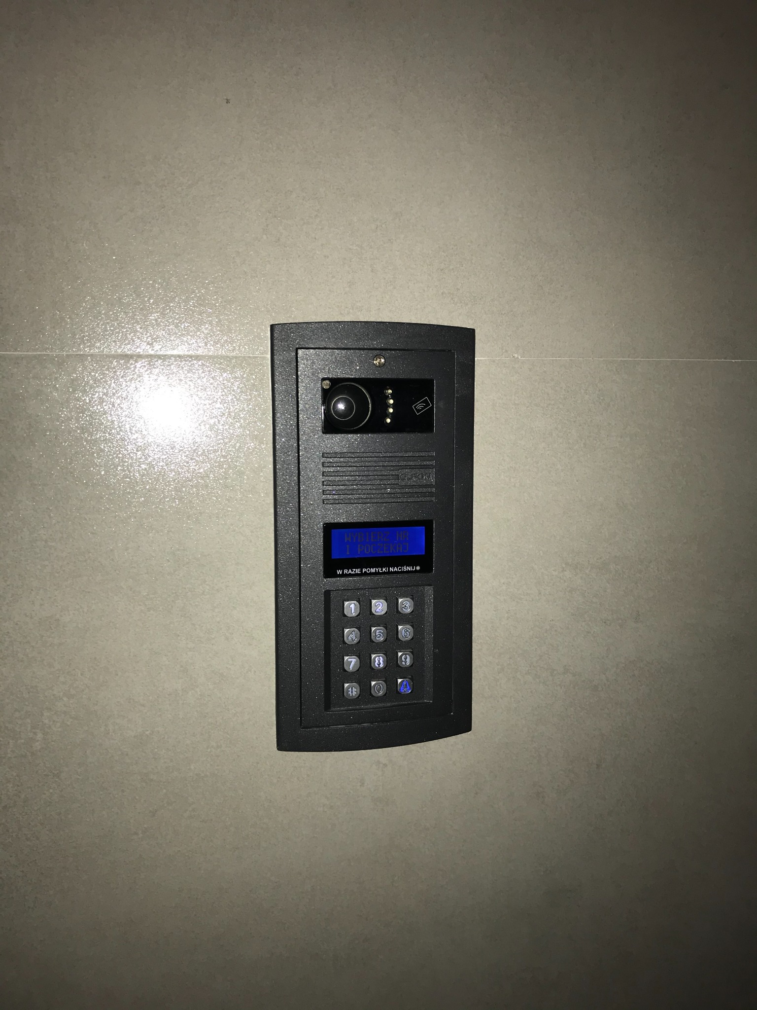 Zamontowany, czarny wideodomofon z kamerą i panelem numerycznym na jasnej, błyszczącej ścianie w Warszawie. Widoczny wyświetlacz z instrukcjami i przyciski numeryczne.