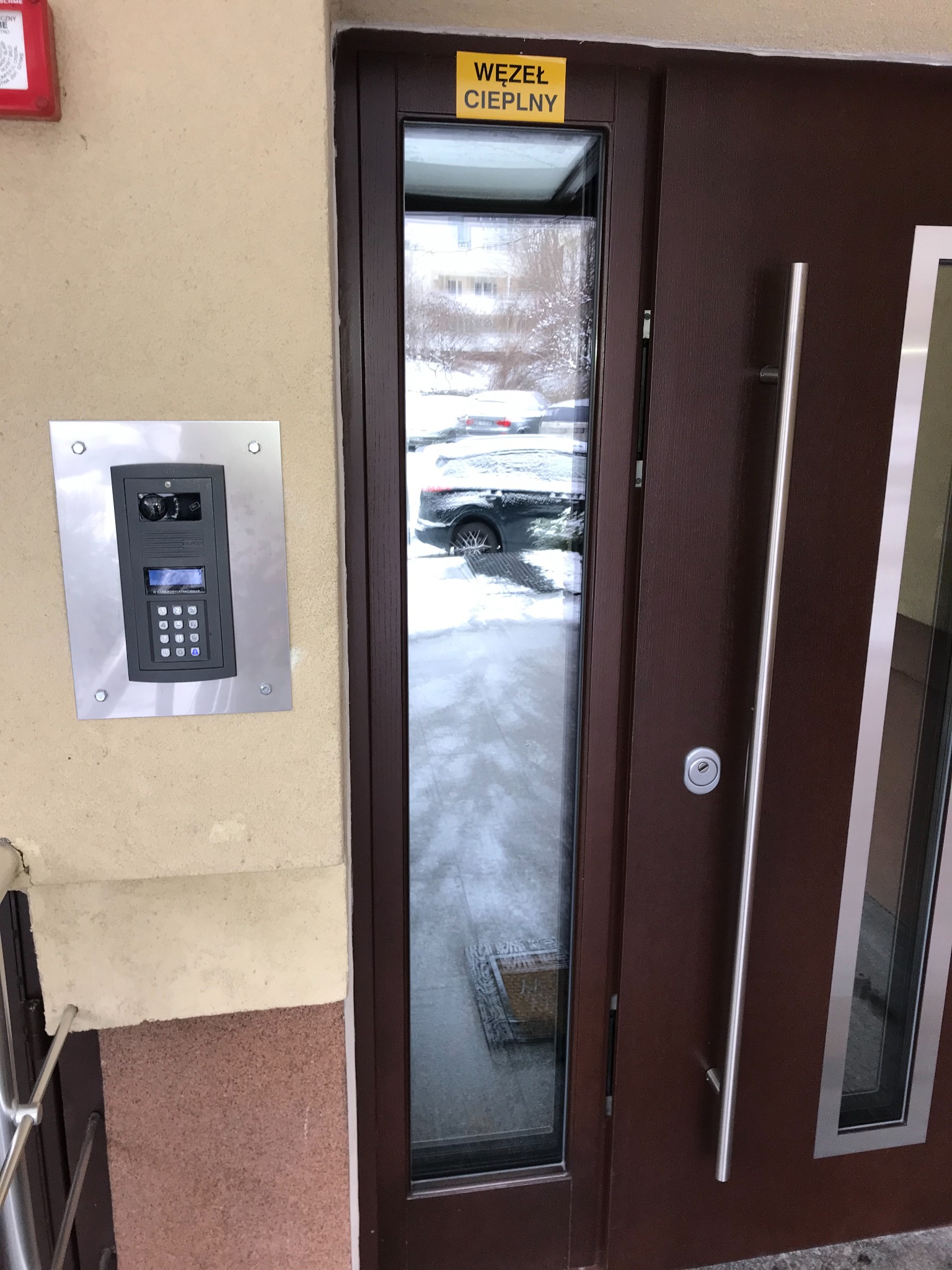 Zewnętrzny panel domofonowy z klawiaturą numeryczną i kamerą, zamontowany obok brązowych drzwi wejściowych z długim, pionowym uchwytem. W tle widok na zasnieżony parking przez szybę w drzwiach.