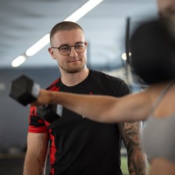 Fotografia i wideografia reklamowa Szymon Kryszczak - Trener personalny w okularach obserwuje kobietę ćwiczącą z hantlą w siłowni, widoczne tatuaże na ramionach obojga.