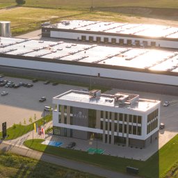 Fotografia i wideografia reklamowa Szymon Kryszczak - Zdjęcie z lotu ptaka przedstawia kompleks budynków przemysłowych z parkingiem pełnym samochodów, w tle pola uprawne, a na budynkach widoczne logo firmy.