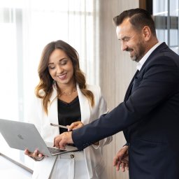 Fotografia i wideografia reklamowa Szymon Kryszczak - Uśmiechnięta kobieta w białym garniturze trzyma laptopa, mężczyzna w granatowym garniturze pokazuje coś na ekranie długopisem, jasne wnętrze biurowe.