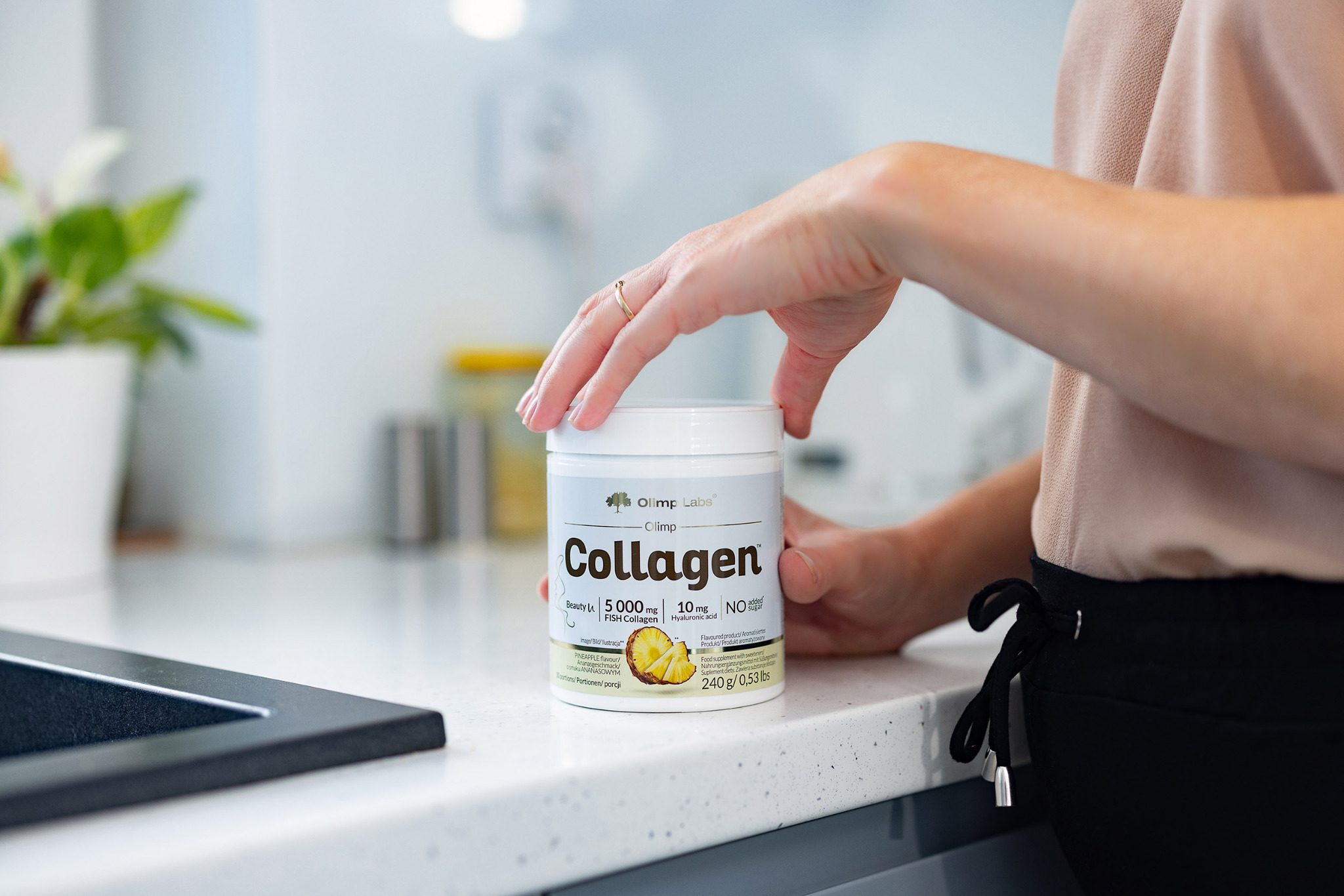 Kobieta otwiera opakowanie kolagenu Olimp Labs na blacie kuchennym z elementami zlewu w tle.