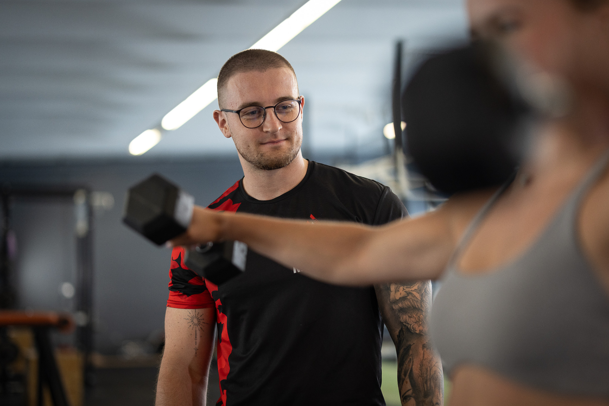 Trener personalny w okularach obserwuje kobietę ćwiczącą z hantlą w siłowni, widoczne tatuaże na ramionach obojga.