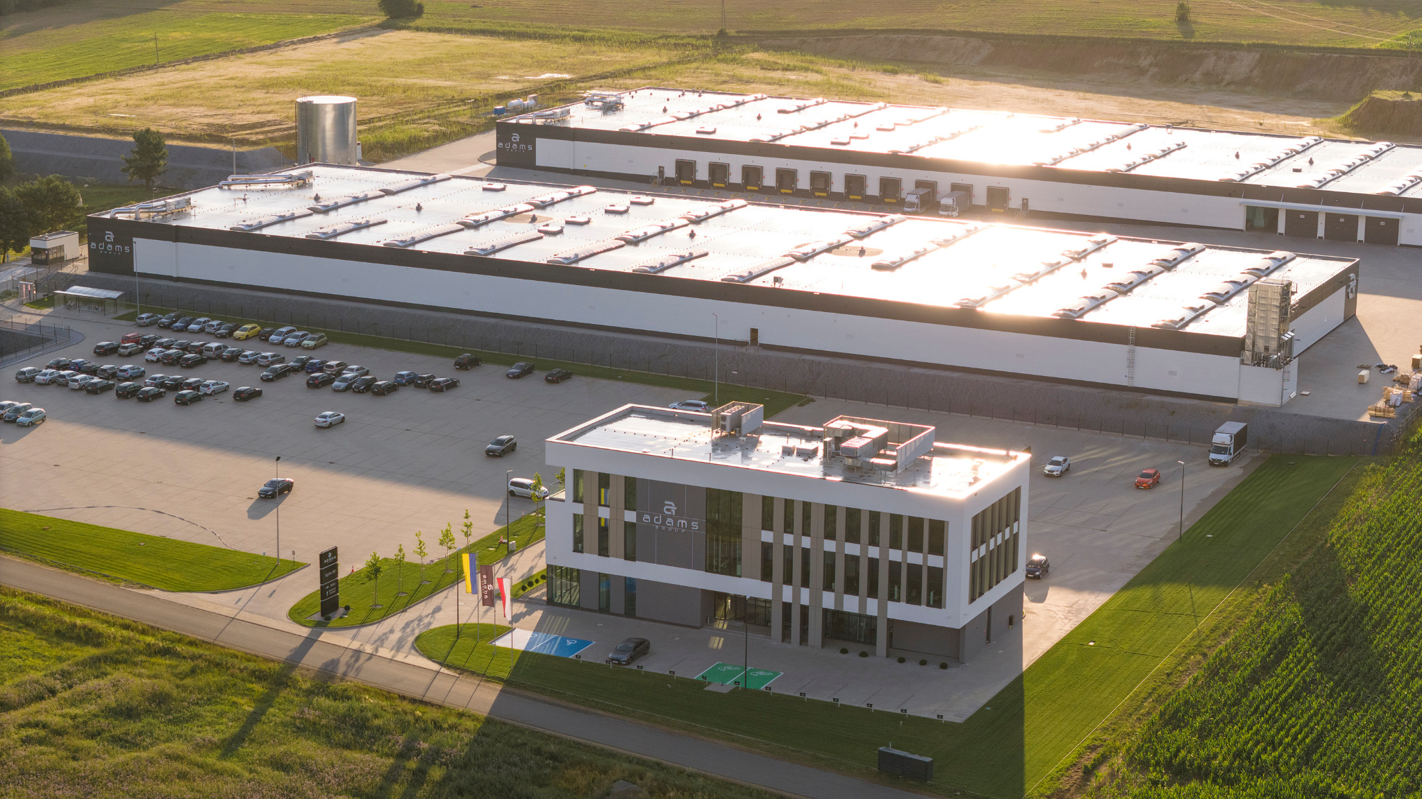 Zdjęcie z lotu ptaka przedstawia kompleks budynków przemysłowych z parkingiem pełnym samochodów, w tle pola uprawne, a na budynkach widoczne logo firmy.