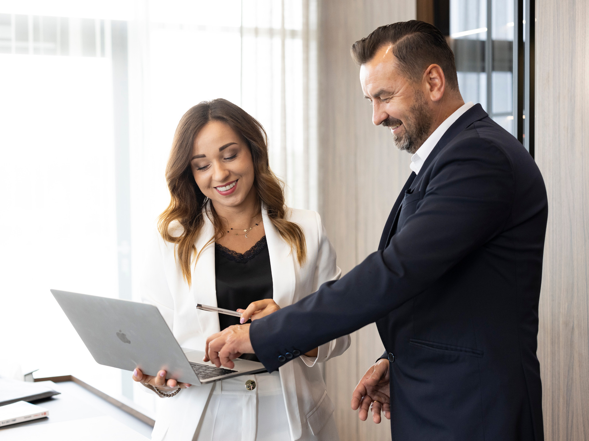 Uśmiechnięta kobieta w białym garniturze trzyma laptopa, mężczyzna w granatowym garniturze pokazuje coś na ekranie długopisem, jasne wnętrze biurowe.