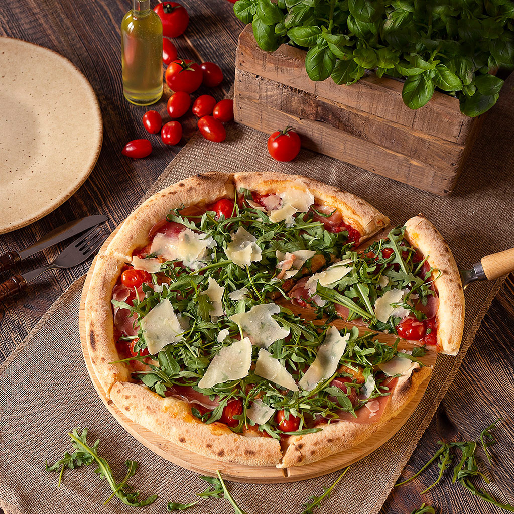 Pizza z rukolą, pomidorkami cherry i parmezanem na drewnianej desce, z wyciętym kawałkiem, w tle bazylia w drewnianej skrzynce, oliwa i talerz.