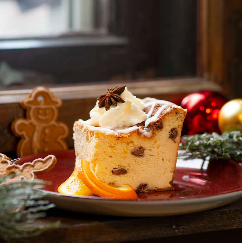 Kawałek sernika z rodzynkami udekorowany bitą śmietaną, anyżem gwiaździstym i plastrami pomarańczy na czerwonym talerzu, w tle pierniczki i świąteczne ozdoby