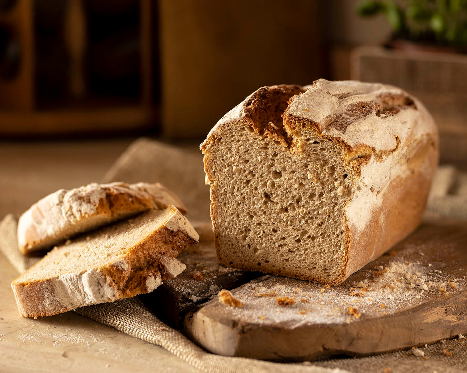 Kromka chleba rustykalnego, przekrojona, z dwoma plastrami obok na drewnianej desce posypanej mąką, w ciepłym oświetleniu.