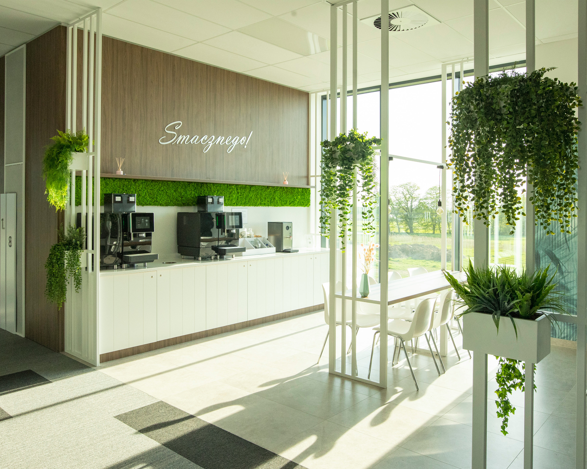 Jasne wnętrze biurowe z ekspresami do kawy, zieloną ścianą z mchu i wiszącymi roślinami. Duże okno z widokiem na zewnątrz. Nowoczesny wystrój.