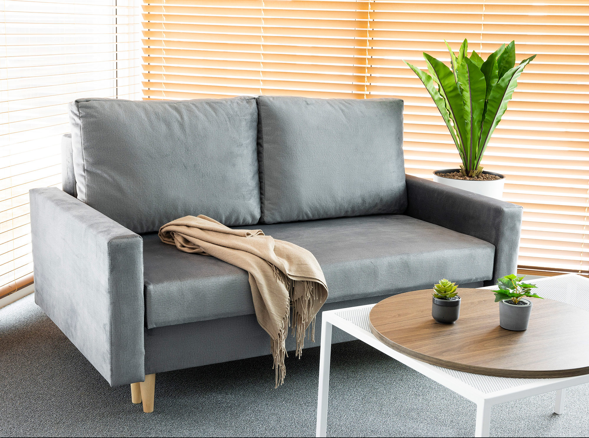 Nowoczesna, szara sofa z beżowym pledem i okrągłym stolikiem kawowym z roślinami w doniczkach na tle drewnianych żaluzji.
