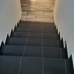 Schody wewnątrz budynku, wyłożone ciemnymi, kwadratowymi płytkami z delikatnym wzorem, prowadzące do pomieszczenia z podłogą z płytek imitujących drewno.