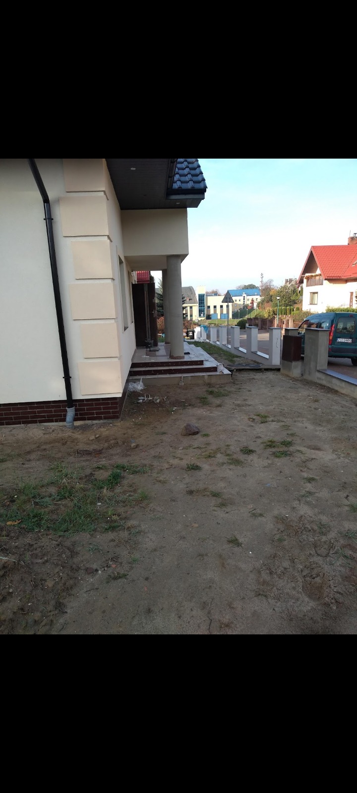 Fragment elewacji domu jednorodzinnego z dekoracyjnymi elementami, widoczne także kolumny przy wejściu i przygotowany teren przed budynkiem.