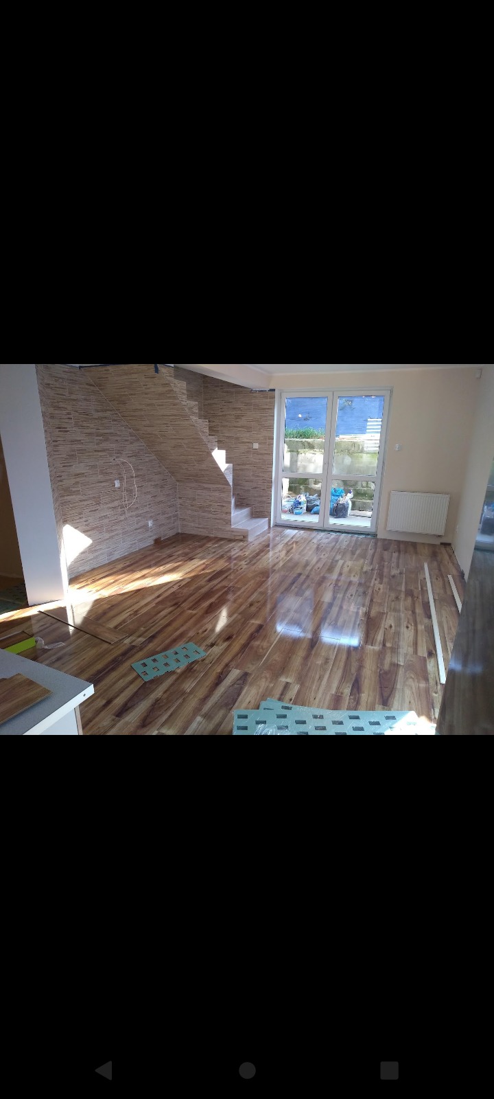 Wnętrze domu po remoncie z nową, błyszczącą podłogą z paneli drewnopodobnych, wykończonymi schodami z płytek imitujących cegłę i widocznymi materiałami izolacyjnymi pod panelami.